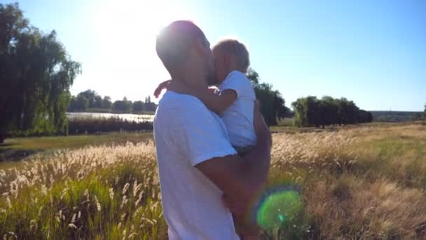Молодий тато тримає дитину на руках на лузі і погладжує його в сонячний день. Батько втішає свого маленького сина на природі. Сім'я проводить час разом. Літній пейзаж на фоні. крупним планом — стокове відео
