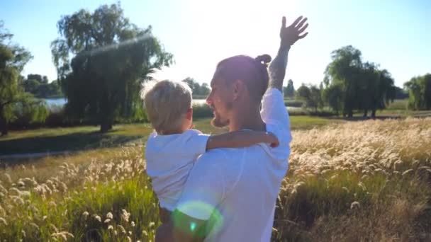 Ojciec i małe dziecko machając rękami na łące. Młody tata trzyma syna na ramionach w przyrodzie. Szczęśliwego spędzania czasu rodzinnego razem. Letni krajobraz w tle. Zamknij tylny widok z tyłu — Wideo stockowe
