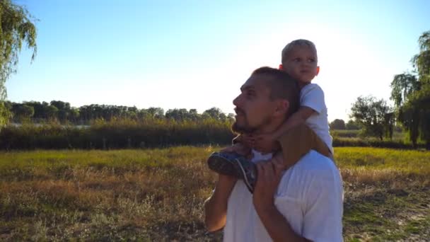 Feliz familia caminando por el prado y relajándose en la naturaleza. Niño con el pelo rubio sentado en los hombros de su padre. Padre e hijo pasando tiempo juntos al aire libre. Luz del sol en el fondo. De cerca. — Vídeo de stock