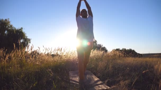 Joven deportista de pie en el yoga posan en la esterilla en el campo en un día soleado. Un tipo caucásico haciendo ejercicio en el prado. Luz del sol en el fondo. Concepto de estilo de vida activo saludable. De cerca. — Vídeos de Stock