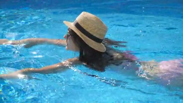 Menina bonita nova em óculos de sol e chapéu nadando na piscina. Mulher relaxando em água morna clara no dia ensolarado. Conceito de férias de verão ou férias. Fechar câmera lenta — Vídeo de Stock