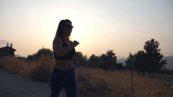 Sportliches Mädchen joggt bei Sonnenuntergang auf einer Landstraße. Junge Frau läuft ins Freie. Gesunder aktiver Lebensstil. Zeitlupe Nahaufnahme — Stockvideo