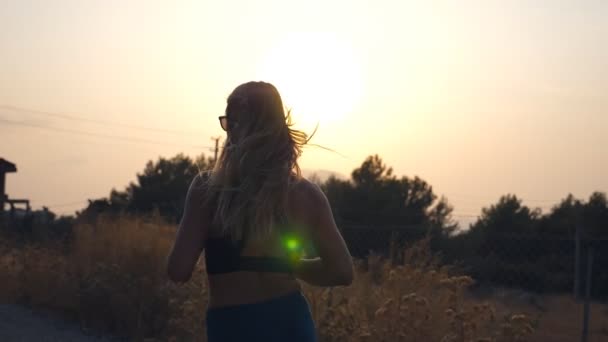 Följ med till sportig tjej och jogga på landsvägen. Ung kvinna i solglasögon springer utomhus och lyssnar musik från smartphone. En hälsosam aktiv livsstil. Slow motion Bakifrån — Stockvideo