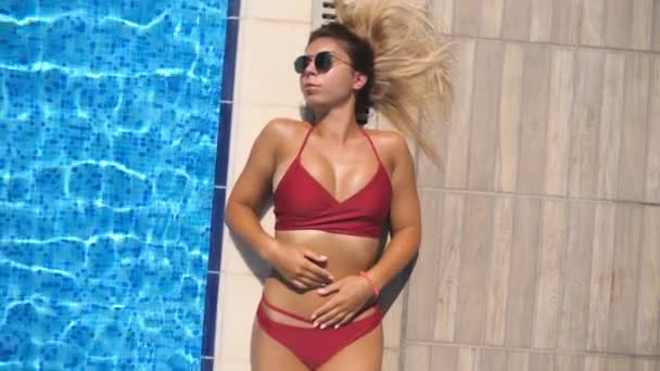 Top view of young tanned girl in sunglasses lying near swimming pool of hotel and touching water surface with her hand. Beautiful woman in red bikini relaxing on the basin edge on sunny day. Close up — Stock Video