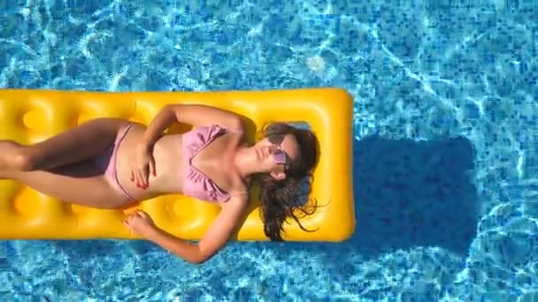 Vista superior da menina bronzeada bonita em óculos de sol e biquíni rosa deitado no colchão inflável amarelo na piscina. Jovem relaxante na bacia do hotel durante as viagens de verão. Conceito de férias — Vídeo de Stock