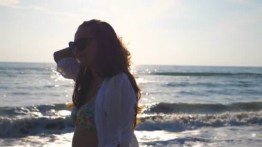 Güzel kız bikini ve sahil yürüyüş ve saçları ile oynarken gömlek portresi. Mutlu genç kadın gülümseyerek ve yaz tatili hayattan zevk güneş gözlüğü. Ağır çekim yakın çekim