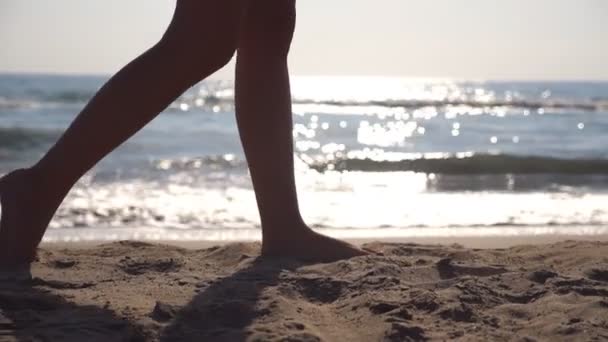 Ženské nohy chůzi na pláži u moře za slunečného dne se vlny na pozadí. Nohy mladé ženy krokování v písku. Letní dovolenou nebo dovolenou koncept. Boční pohled zpomalené Detailní záběr — Stock video
