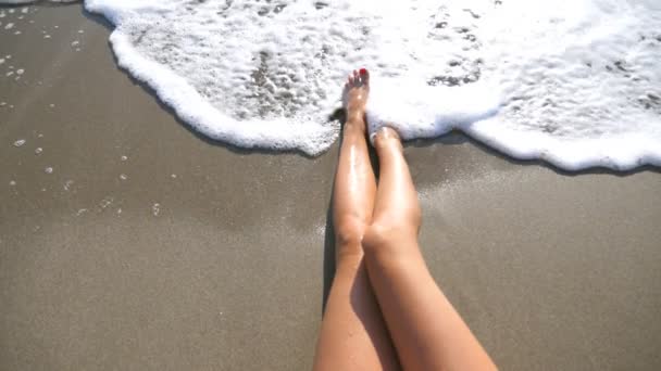 海の波は、日焼けした女性の足を洗います。美しい少女は、晴れた日にビーチで横になっています。夏休みや休日中に海岸で若い女性がリラックスできます。上から見るスローモーションをクローズ アップ — ストック動画