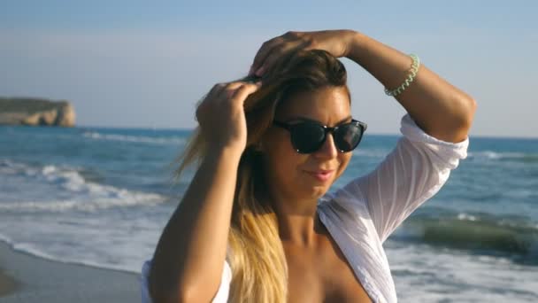 Close up of beautiful girl in bikini and shirt walking along shore and playing with her blonde hair on sunny day. Happy young woman in sunglasses smiling and enjoying summer vacation on beach. Slow mo — Stock Video