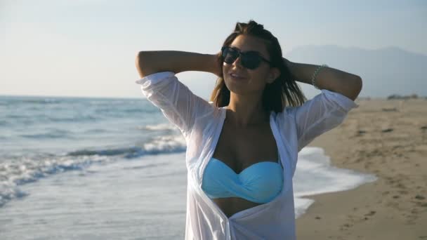 Menina bonita em biquíni e camisa andando ao longo da costa e tocando seu cabelo loiro. Jovem feliz em óculos de sol sorrindo e curtindo a vida durante as férias. Conceito de férias de verão. Movimento lento — Vídeo de Stock