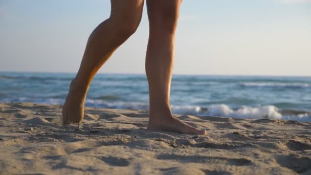 Pés femininos andando descalços na praia durante o dia ensolarado. Pernas de uma jovem a pisar na areia. Menina descalça na costa. Ondas no fundo. Conceito de férias de verão ou férias. Fechar câmera lenta — Vídeo de Stock