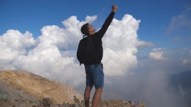 Sırt çantasıyla dağın tepesine uzanan ve elini kaldıran genç bir erkek yürüyüşçü. Çocuk turist tepenin kenarında duruyor ve muzaffer bir şekilde kolunu uzatıyor. Özgürlüğün tadını çıkaran adam. Yavaş çekim