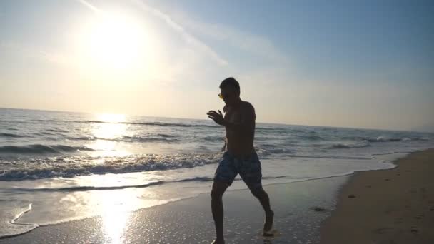 Sylwetka młodego sportowca działa szybko wzdłuż wybrzeża podczas wschodu słońca. Sportowe szkolenie chłopca na plaży z fal morskich w tle. Mężczyzna sportowca ćwiczenia na świeżym powietrzu. Zdrowy styl życia. Zwolnionym — Wideo stockowe