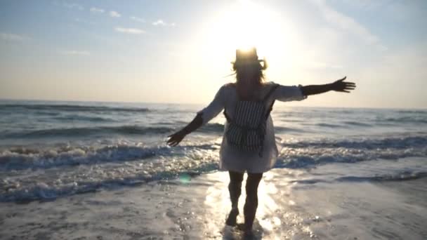 Jovem mulher feliz de camisa com mochila correndo para o mar e pulverizando água com os pés ao nascer do sol. Menina bonita em chapéu desfrutando de férias de verão ou férias. Conceito de viagem Visão traseira Movimento lento — Vídeo de Stock