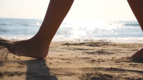 Close-up van vrouwelijke voeten lopen op gouden zand op het strand met oceaan golven op de achtergrond. Poten van een jonge vrouw die in het zand stapt. Blootsvoets meisje aan de kust. Zomervakantie. Langzame beweging — Stockvideo