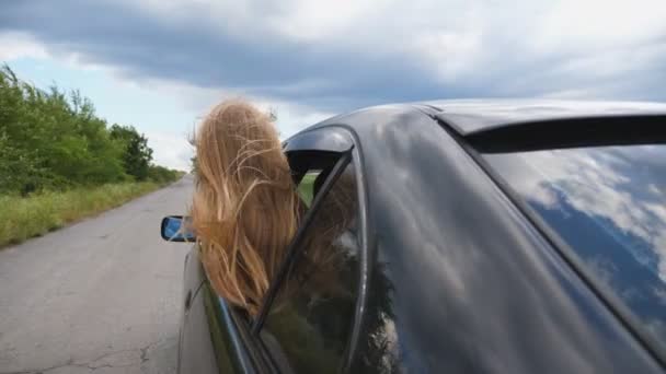 Little kid with long blonde hair leaning out of car window and enjoying road trip. Beautiful small girl looking out of open window moving auto while riding through country road at overcast weather — Stock Video