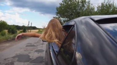 Küçük kız araba penceresinden dışarı eğilerek ve kırsal yolda sürüş sırasında rüzgar elini sallayarak. Küçük çocuk açık pencereden dışarı bakarak otomatik hareket ve kolu esinti ile oynuyor. Arka görüş