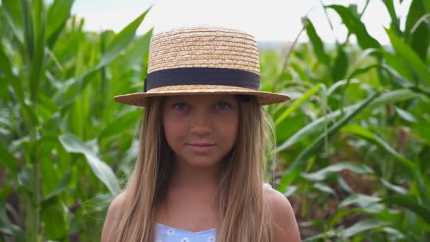 Primer plano de la niña sonriente en sombrero de paja mirando a la cámara en el prado en la granja orgánica. Retrato de un niño pequeño y feliz con el pelo largo y rubio de pie sobre el fondo borroso del campo de maíz — Vídeo de stock