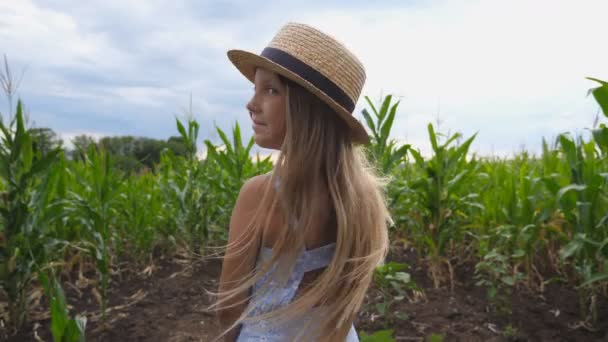 Attraente bambina in cappello di paglia in piedi al campo di mais girando verso la fotocamera e sorridendo. Piccolo bambino con lunghi capelli biondi che guarda piantagione di mais e godersi il paesaggio naturale. Chiudere Lento mo — Video Stock