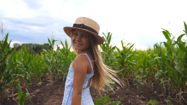 Close-up van mooi klein meisje in stro hoed staande op maïsveld, wenden zich tot de camera en glimlachend. Kleine jongen met lange blonde haren kijken naar maïs plantage en genieten van natuur landschap. Slow mo — Stockvideo