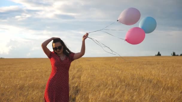 Joyeux belle fille dans des lunettes de soleil marchant à travers le champ de blé et tenant des ballons à la main. Jeune femme en robe rouge allant parmi la plantation d'orge et redressant ses longs cheveux bruns. Mouvement lent — Video