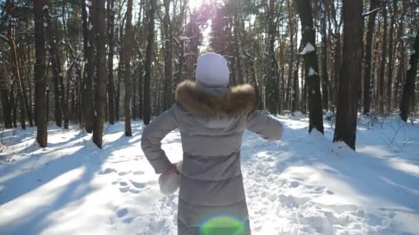 Widok z tyłu młodej kobiety spacerującej w słonecznym lesie zimowym. Nierozpoznawalna dziewczyna ciesząca się przechadzką po śnieżnym lesie podnosząc ręce. Beztroska pani bawiąca się na świeżym powietrzu. Piękny krajobraz. Zwolniony ruch — Wideo stockowe