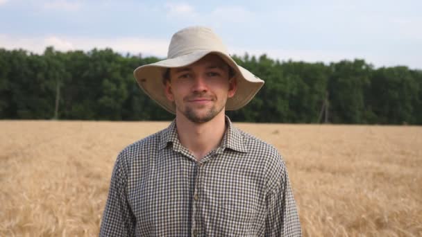 Detailní záběr šťastného usměvavého farmáře v klobouku, jak se dívá do kamery na rozmazaném pozadí pšeničného pole. Portrét pohledného mladého agronoma stojícího na ječné louce. Koncept zemědělství — Stock video