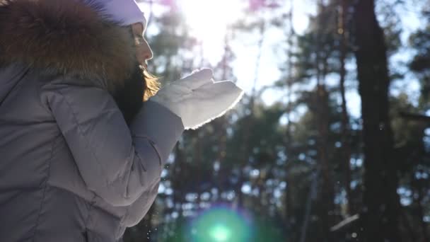Typ av vacker kvinna i vita handskar som blåser av snö från handflatorna. Ung brunett flicka som står bland vinterskog och leker med snö på solig dag. Ljus sol skiner i bakgrunden. Långsam rörelse — Stockvideo