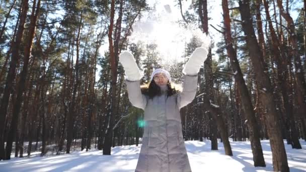 Молода щаслива жінка в зимовому одязі кидає жменьку снігу і насолоджується падаючими сніжинками. Мила дівчина грає зі снігом в прекрасний зимовий день. Мальовничий засніжений ліс на фоні. Повільний рух — стокове відео