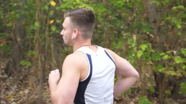 Giovane sportivo che corre lungo il sentiero nei pressi della foresta all'inizio dell'autunno. Bello sportivo con cuffie wireless che fanno jogging all'aperto. Un atleta che fa ginnastica nella natura. Stile di vita sano e attivo. Vista laterale — Video Stock