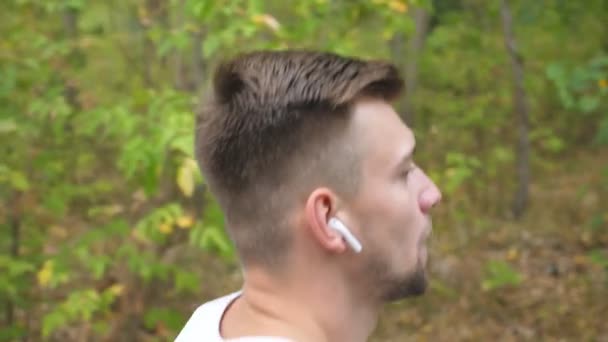 Close up face of young strong sportsman sprinting fast along trail near forest. Athletic man with wireless headphones jogging at early autumn. Handsome guy running at nature. Healthy active lifestyle — Stock Video