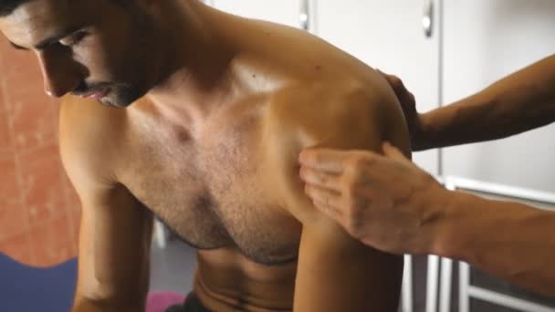 Beau sportif musclé assis sur la table de massage et les bras du masseur massant son épaule dans le salon. Les mains masculines du masseur professionnel faisant frottement de guérison au jeune athlète dans le salon. Gros plan — Video