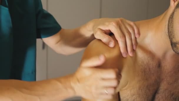 Închideți mâinile masculine ale masagistului profesionist de vindecare a umărului de masaj al sportivului muscular care stă pe masa de masaj în salon. Brațele de maseur face frecare la atlet tânăr în salon. Slow motion — Videoclip de stoc