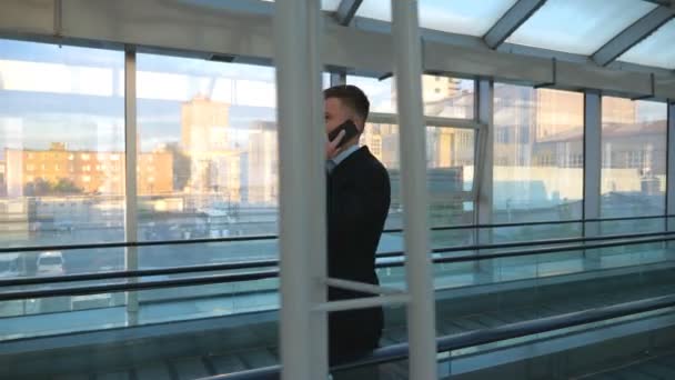 Pewny siebie biznesmen przechodzący przez szklaną halę terminalu z bagażem i rozmawiający przez telefon. Odnoszący sukcesy przedsiębiorca rozmawiający o pracy w drodze na lot. Powolny ruch Widok z boku — Wideo stockowe