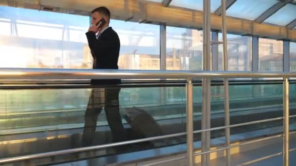Kendine güvenen işadamı, cam terminalde bavuluyla yürüyor ve telefonla konuşuyor. Başarılı bir girişimci uçuşa hazırlanırken iş görüşmesi yapıyor. Yavaş çekim kenar görünümü — Stok video