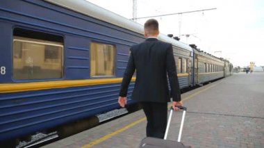 Siyah takım elbiseli tanınmayacak kadar başarılı bir işadamı platformda yürüyor ve tekerlekli bavulu çekiyor. Kendinden emin genç bir adam, bavuluyla trenin yanından geçiyor. Yavaş çekim Arka plan