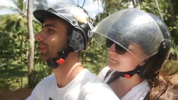 Happy young couple in helmets rides an motor scooter along country road. Smiling man and woman drives a moped along driveway on a tropical island. Summer vacation or holiday concept. POV Slow motion — Stock Video