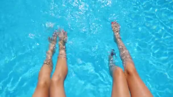 Jonge vrouwen die op de rand van het zwembad zitten en hun voeten in het water bungelen. Twee meisjes met slanke gebruinde benen ontspannen in de buurt van het bassin. Dames genieten van vakantie in het resort. Begrip zomervakantie. Bovenaanzicht — Stockvideo