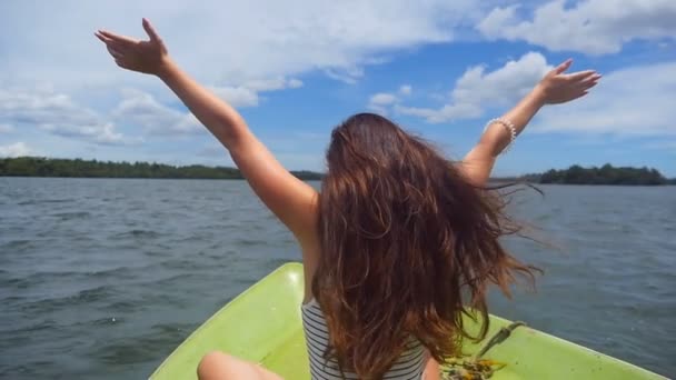 Young woman with blowing brown hair sits on bow of boat and raises hands. Unrecognizable girl relaxes on deck of ship and admires a beautiful nature landscape. Vacation or holiday concept. Dolly shot — Stock Video