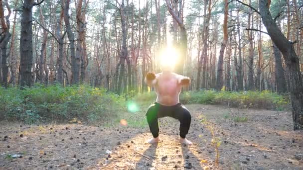 Genç adam güneşli bir ormanda çalışıyor. Sportif adam gün batımında çömelme hareketleri yapıyor. Doğada atlet eğitimi. Sağlıklı ve aktif yaşam tarzı kavramı. Dolly vuruldu. Yaklaş. — Stok video