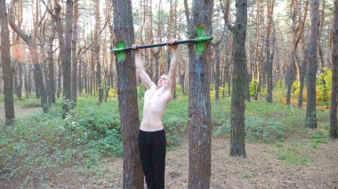 Güçlü ve kaslı bir adam güzel ormanda barfiks çekiyor. Erkek sporcu doğada yatay çubukta egzersiz yapıyor. Atletik adam dışarıda antrenman yapıyor. Spor anlayışı ve aktif yaşam tarzı. Dolly vurdu..