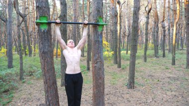 Güçlü ve kaslı bir adam güzel ormanda barfiks çekiyor. Erkek sporcu doğada yatay çubukta egzersiz yapıyor. Atletik adam dışarıda antrenman yapıyor. Spor anlayışı ve aktif yaşam tarzı. Yavaş çekim.