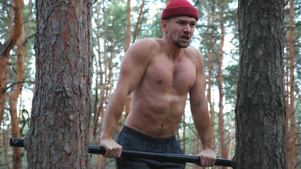 Strong and muscular man doing push ups on horizontal bar in forest. Hardy sportsman working out at nature. Athletic guy training outdoor. Concept of sport and active lifestyle. Close up Slow motion.