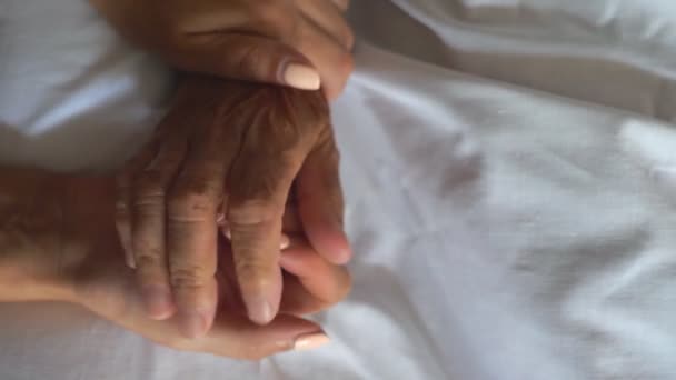Oigenkännlig kvinna som håller hand med sin gamla mor som ligger i sängen och visar omsorg eller kärlek. Dottern rör försiktigt skrynkla arm äldre mamma. Barnbarn smeker mormor hand ger stöd. Ovanifrån — Stockvideo