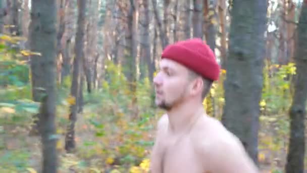Kırmızı şapkalı güçlü ve kaslı bir adam orman yolunda koşuyor. Erkek sporcu, dışarıda çalışıyor. Kardiyo antrenmanı yapan cesur bir sporcu tahtada koşuyor. Sağlıklı aktif yaşam tarzı kavramı — Stok video