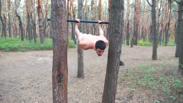 Silny mężczyzna pokazuje gimnastykę na poziomym drążku. Muskularny facet ćwiczący elementy gimnastyczne. Sportowiec wykonuje ćwiczenia siłowe podczas treningu w lesie. Trening sportowy ćwiczenia statyczne — Wideo stockowe