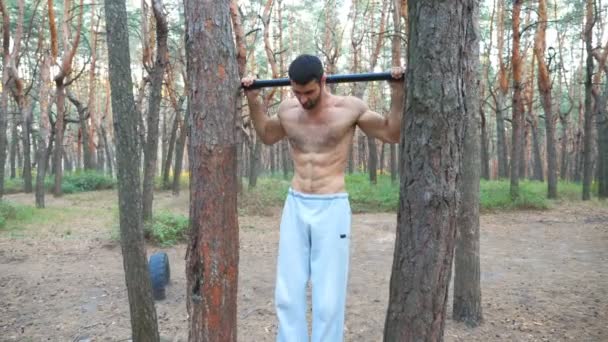 Silny mężczyzna pokazuje niektóre ćwiczenia na belce poziomej. Muskularny facet podciąga się. Sportowiec wykonuje trening siłowy w lesie. Trening sportowy na świeżym powietrzu. Pojęcie sportu i aktywnego stylu życia — Wideo stockowe