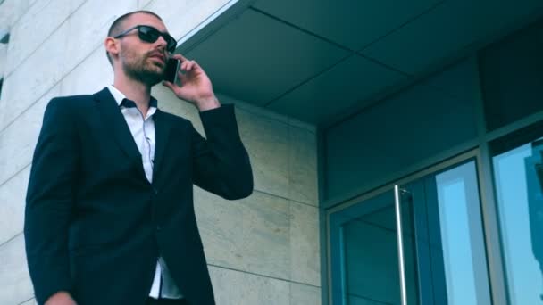 Businessman in sunglasses walking near office and talking on phone. Young happy business man going outdoor and speaking on cellphone. Portrait of guy having work conversation. Slow motion Close up — Stock Video