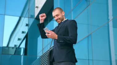 Genç işadamı ofis yakınında akıllı telefon kullanıyor ve başarıyı kutluyor. İş adamı telefonda iyi haberleri ve olumlu duyguları öğrendi. Erkek portresi başarıyla mutludur. Yavaş çekim