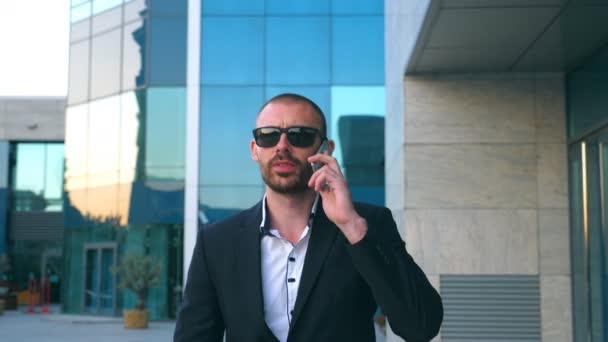 Portrait of businessman in sunglasses talking on phone and walking in street. Young man having business conversation during commuting to work. Confident guy in suit being on his way to office building — Stock Video